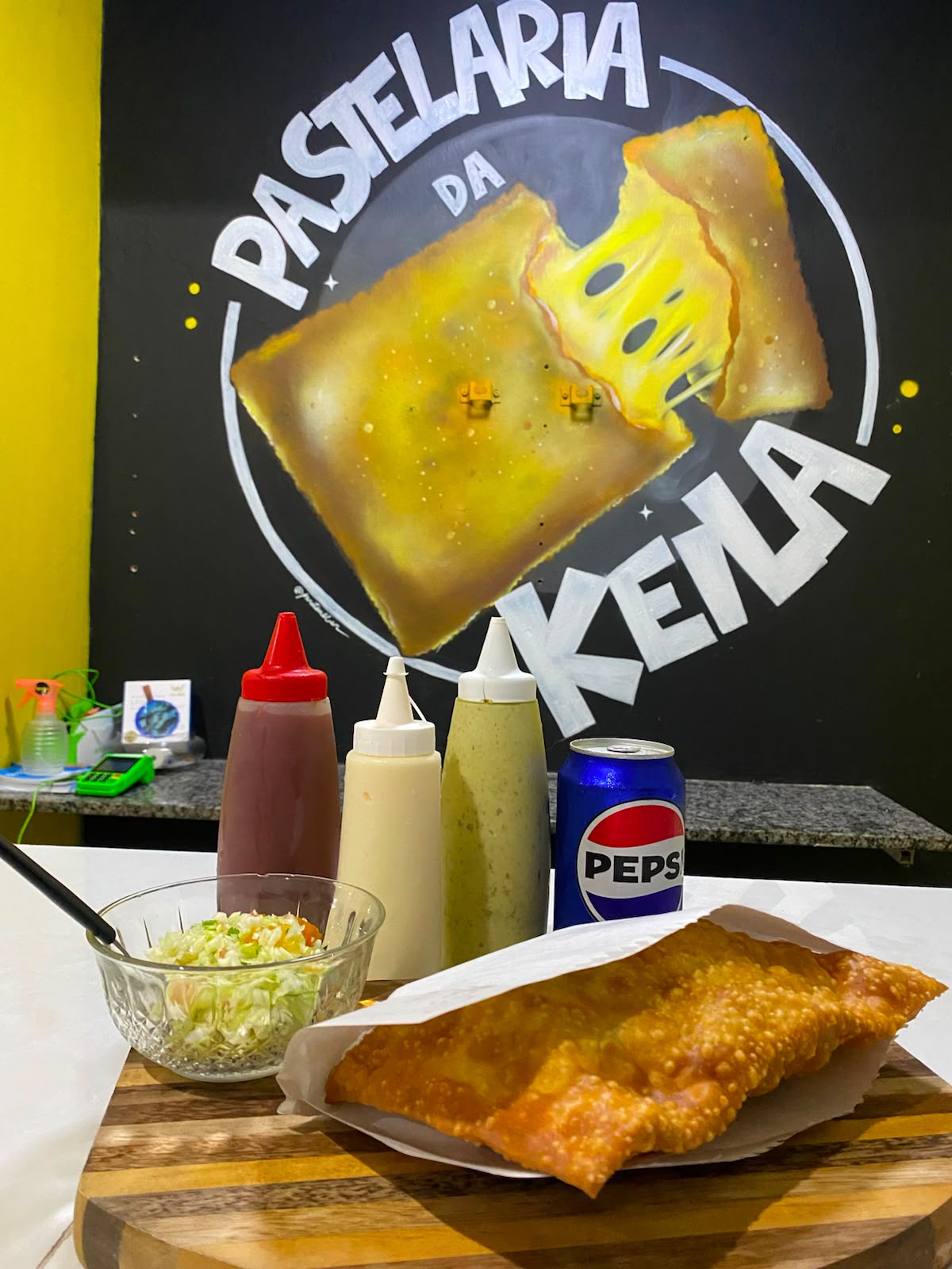 Cantinho da Pastelaria da Keila, com o mural da marca, molhos e um pastel fresquinho